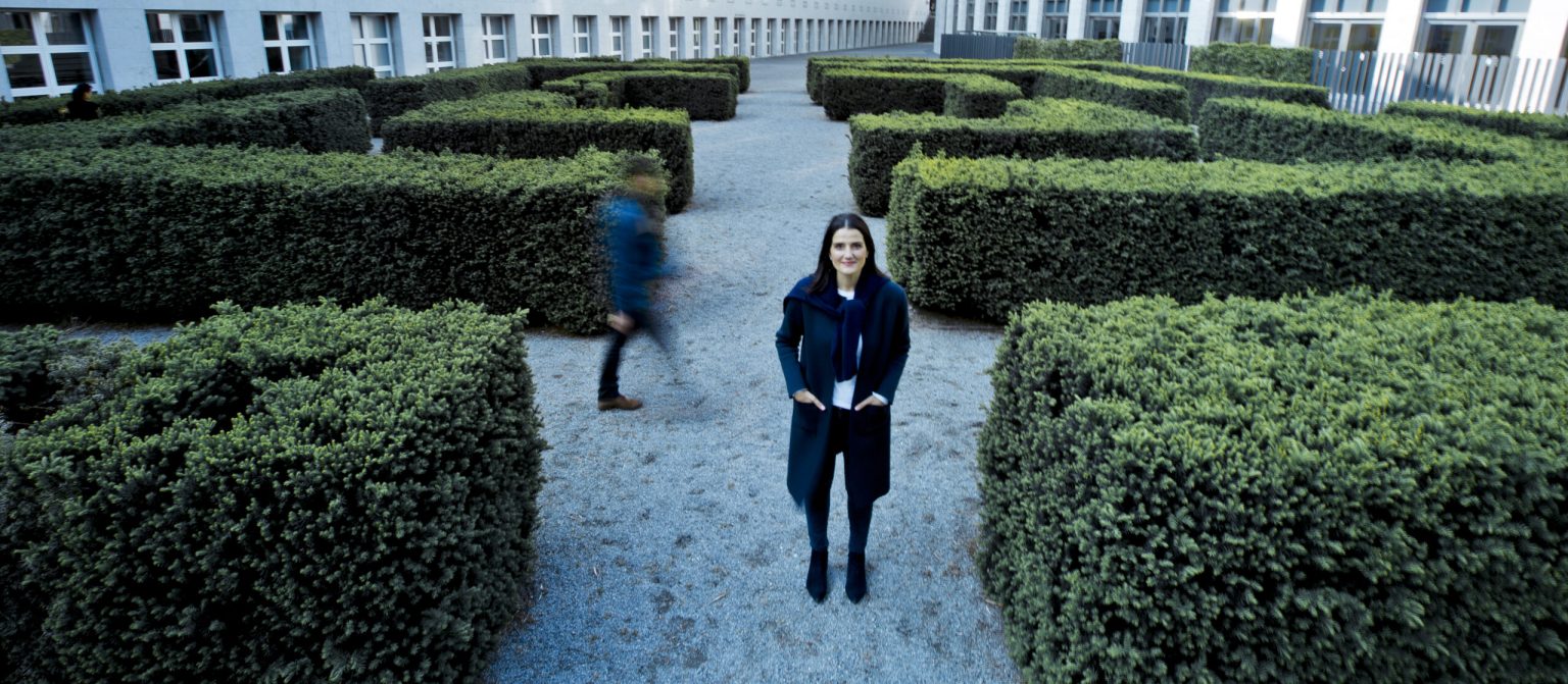 Irena Mila Steinmeier steht am Ausgang eines Heckenlabyrinths, eine Person geht vorbei – Coaching für Geschäftspartner